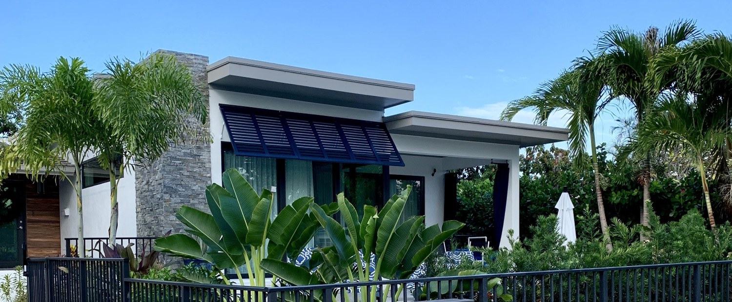 The finished addition from the side, with the stone column, navy metal awning, deep roof overhang and mature palms
