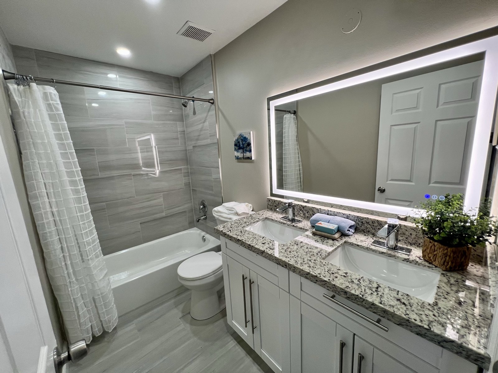 Completed guest bathroom with a double granite vanity, backlit mirror, tiled tub surround and tile flooring