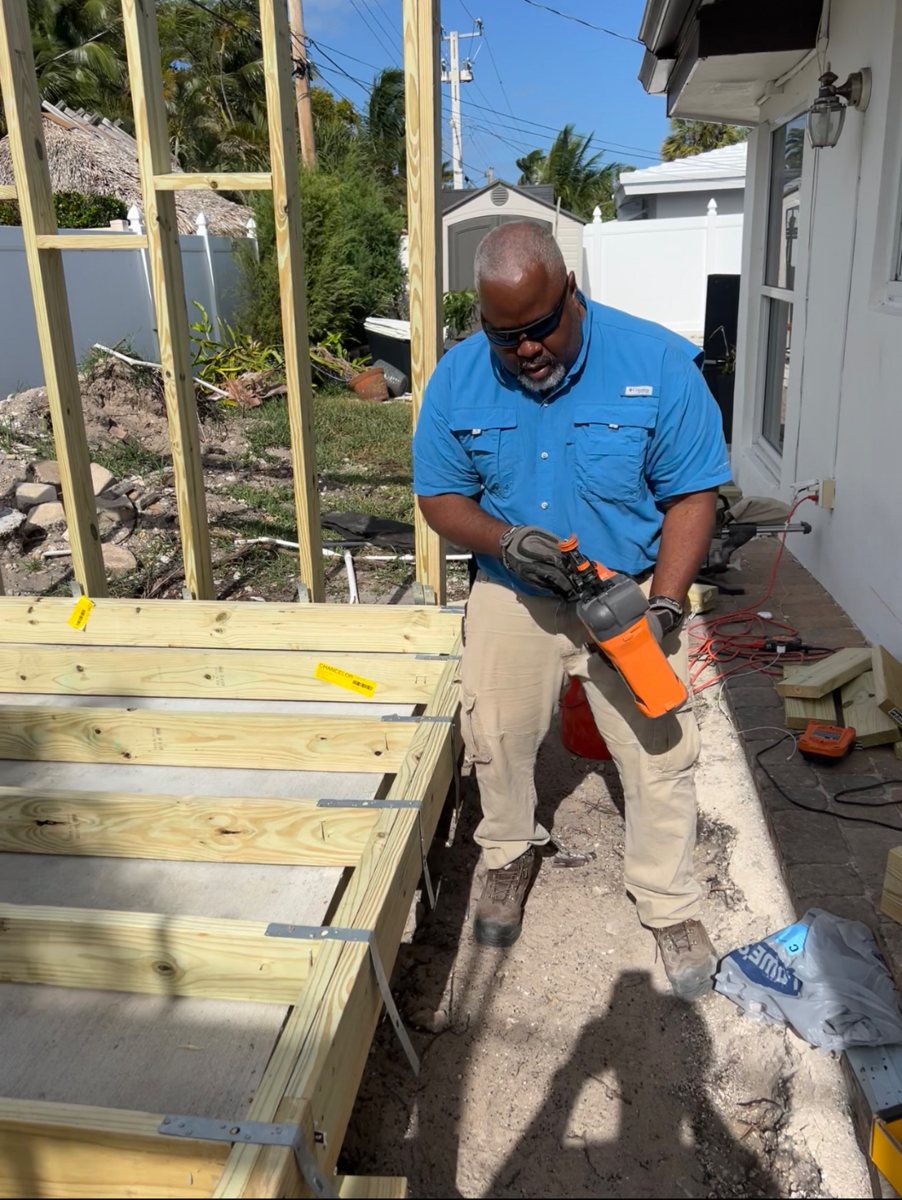 Framing a pressure-treated deck structure with a nail gun, joists set in galvanized hangers alongside the house
